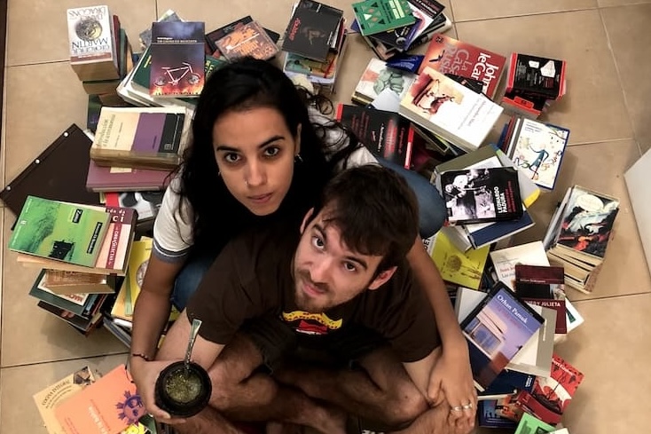 Two people from above between a bunch of books on the floor looking up to the camera